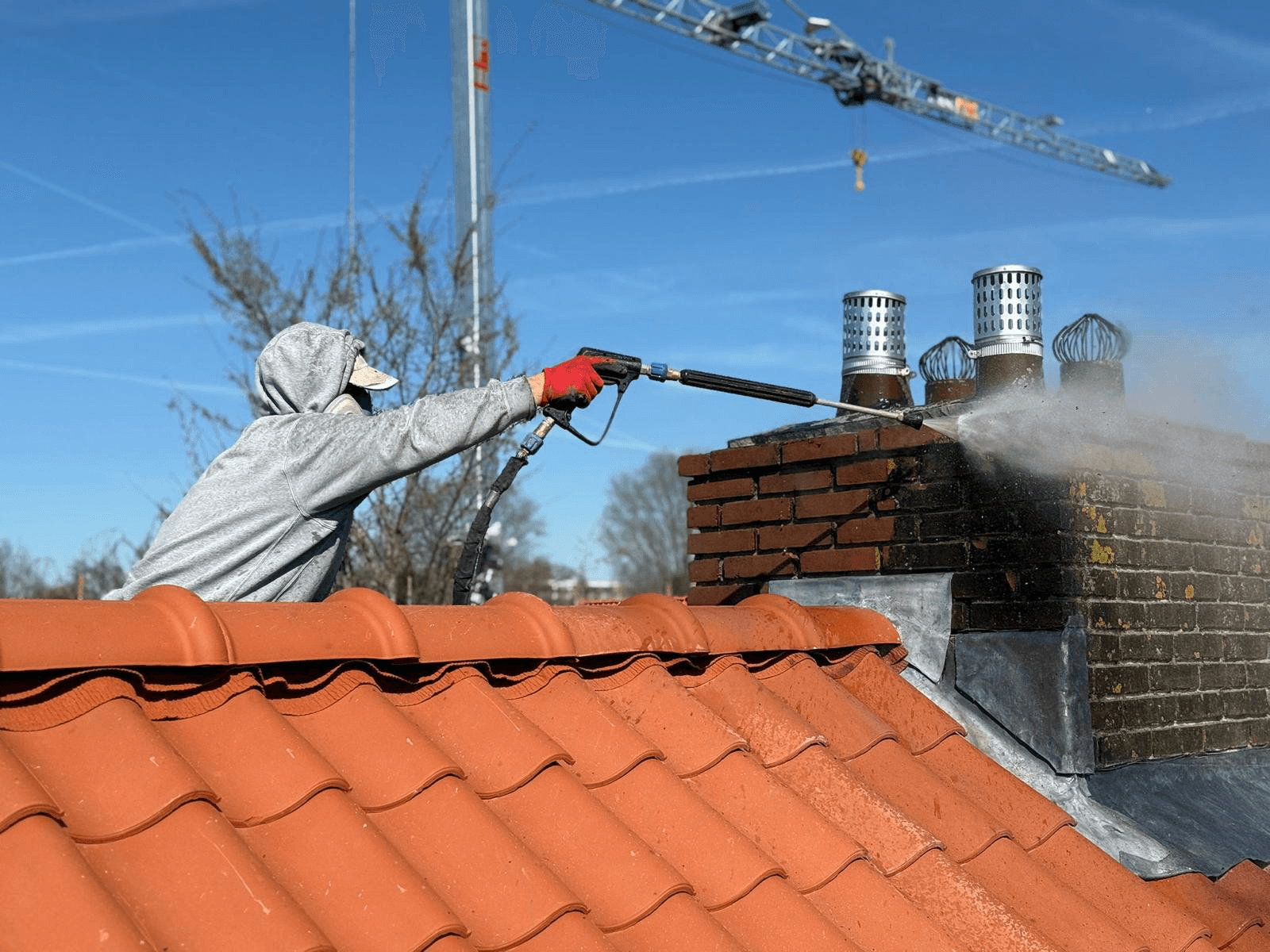 Renovox medewerker stoomt schoorsteen schoon op terracotta dak, blauwe lucht met bouwkraan